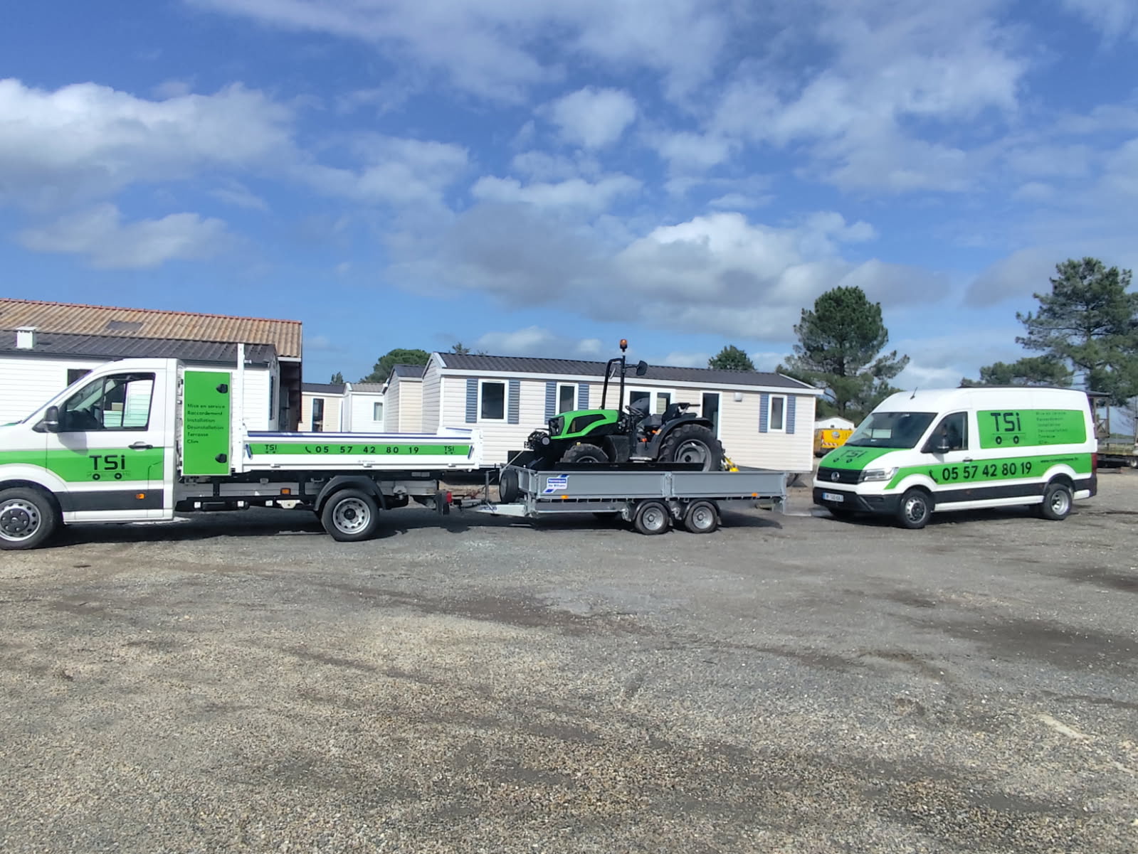 Véhicules utilitaires de l'entreprise TSi avec remorque transportant un tracteur d'installation devant des mobil-homes sous un ciel bleu.